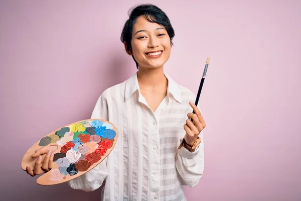 若い美しい中国の芸術家の女性は 歯を示す自信を持って笑顔で立って笑顔で幸せな顔と色の絵筆とパレットを使用して絵を描く — ストック写真