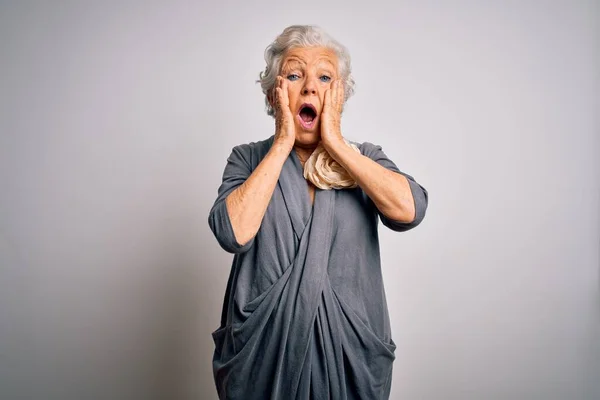 Senior beautiful grey-haired woman wearing casual dress standing over white background afraid and shocked, surprise and amazed expression with hands on face