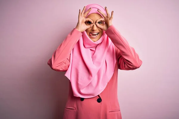 ピンクのイスラム教徒のヒジャーブを身に着けている若い美しいブルネットのビジネスマンの女性とビジネスジャケットは 双眼鏡が舌を突き出すようなOkジェスチャーをして 目は指を通して見ている 狂気の表現 — ストック写真