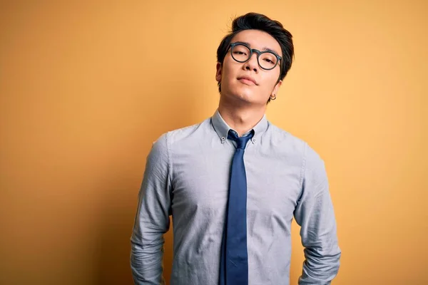Young handsome chinese businessman wearing glasses and tie over yellow background Relaxed with serious expression on face. Simple and natural looking at the camera.