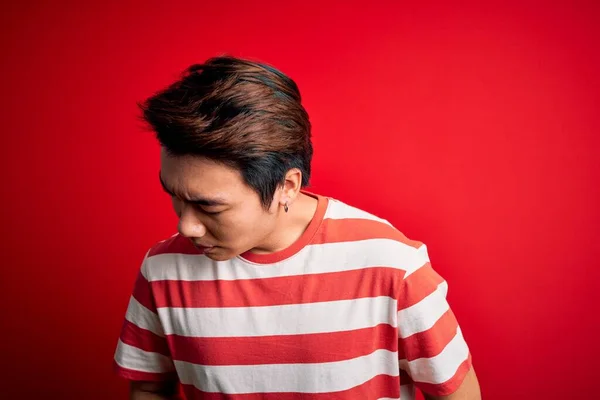 若いハンサムな中国人男性が胃の上に手で赤い背景に立ってカジュアルな縞模様のTシャツを着て消化不良 痛みを伴う病気の気分が悪い アチェの概念 — ストック写真
