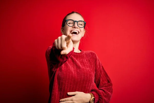 Young beautiful redhead woman wearing casual sweater over isolated red background laughing at you, pointing finger to the camera with hand over body, shame expression