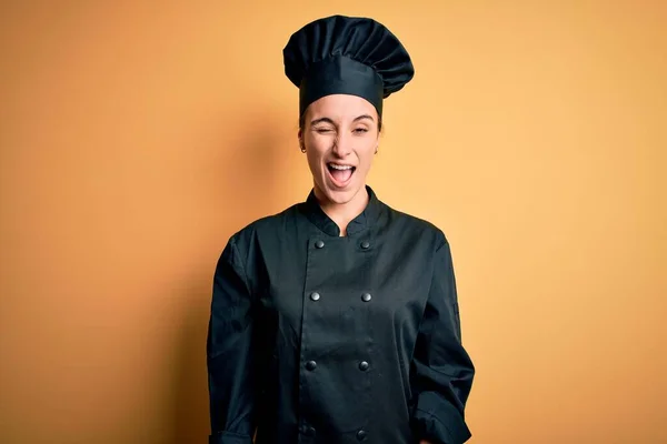 年轻美丽的厨师女士身穿炊事服 头戴一顶帽子 站在黄色的背景上眨眼看着摄像机 脸上洋溢着性感的表情 笑容满面 — 图库照片