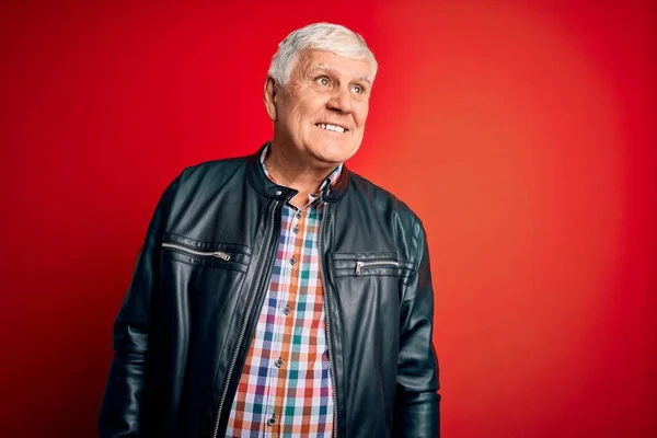 Senior handsome hoary man wearing casual shirt and jacket over isolated red background looking away to side with smile on face, natural expression. Laughing confident.
