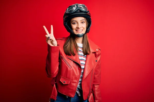 若い美しいブルネットのオートバイのヘルメットと赤いジャケットを身に着けているオートバイの女性が表示され 指番号2で指摘しながら 自信と幸せ笑みを浮かべて — ストック写真