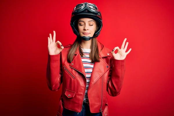 オートバイのヘルメットと赤いジャケットを身に着けている若い美しいブルネットの元サイクリストの女性は 指で瞑想のジェスチャーをして閉じ目でリラックスして笑顔 ヨガのコンセプト — ストック写真