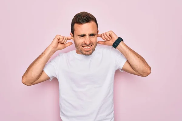 英俊的蓝眼睛男子穿着休闲的白色T恤 站在粉色背景上 用手指捂住耳朵 对嘈杂的音乐感到恼怒 聋人概念 — 图库照片