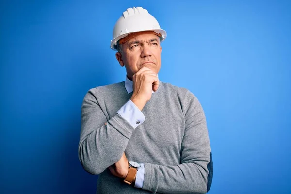 Middle age handsome grey-haired engineer man wearing safety helmet over blue background with hand on chin thinking about question, pensive expression. Smiling with thoughtful face. Doubt concept.