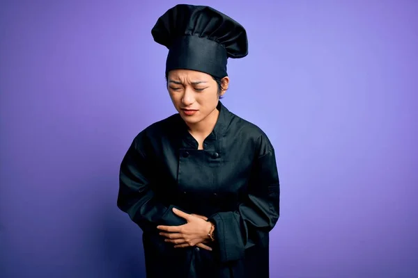 Young beautiful chinese chef woman wearing cooker uniform and hat over purple background with hand on stomach because indigestion, painful illness feeling unwell. Ache concept.