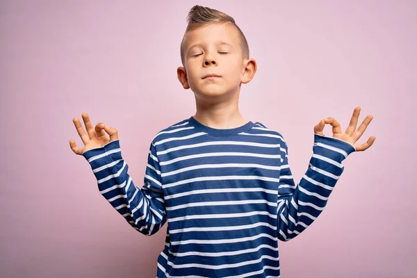 ピンクの背景にネイビーのストライプのシャツを着た青い目の若い白人の子供はリラックスして笑顔で目を閉じて指で瞑想のジェスチャーをしています ヨガのコンセプト — ストック写真