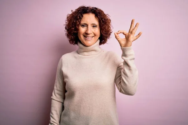 ピンクの背景にカジュアルなタートルネックセーターを身に着けている中世の美しい巻き毛の女性は 手と指でOkサインを行う積極的な笑顔 成功した表現 — ストック写真