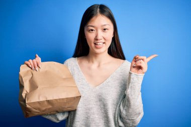 Asyalı genç bir kadın, servis poşeti taşıyor mavi izole edilmiş arka planda yemek için. Elleri ve parmakları kenarı işaret ederek çok mutlu.