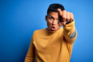 İzole edilmiş mavi arka planda sarı süveter giyen genç, yakışıklı, Latin bir adam. Parmakları önde, ağzı açık, şaşırmış, ön tarafta bir şey var.