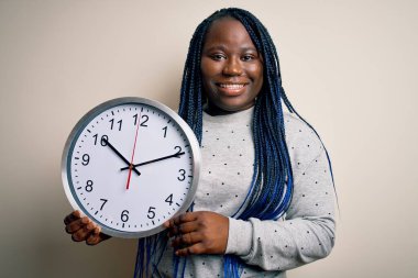 Genç Afro-Amerikalı, saçları örgülü, elinde kocaman bir saat tutan, mutlu yüzüyle gülümseyen, kendine güvenen, dişlerini gösteren iri yarı bir kadın.