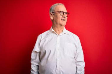Orta yaşlı yakışıklı, beyaz gömlekli, kırmızı arka planda gözlüklü, yüzünde gülümseme ve doğal bir ifadeyle yan yana bakan bir adam. Kendinden emin gülen.