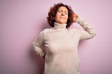 Orta yaşlı güzel kıvırcık saçlı kadın pembe arka planda balıkçı yaka kazak giyiyor boynu ağrıyor, elleriyle boynuna dokunuyor, kas ağrısı çekiyor.