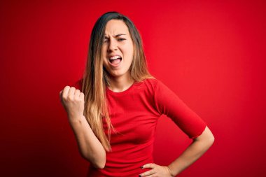 Genç, sarışın, mavi gözlü, kırmızı arka plan üzerine gündelik tişört giyen öfkeli ve öfkeli bir şekilde yumruklarını kaldıran öfkeli bir kadın. Öfke ve agresif kavram.