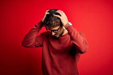 Sakallı, gözlüklü ve süveterli genç adam kırmızı arka planda dikiliyor. Ağrı ve migrenden dolayı çaresiz ve stresli. Ellerini başının üstüne koy..