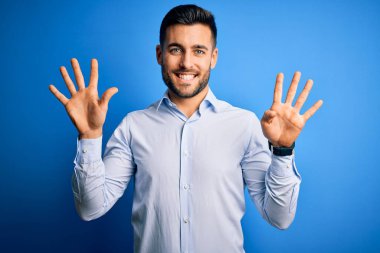 Zarif gömlekli genç adam mavi arka planda dikilip dokuz numaralı parmaklarıyla işaret ederken kendinden emin ve mutlu bir şekilde gülümsüyor..