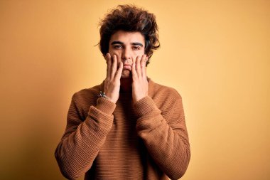 İzole edilmiş sarı arka plan üzerinde gündelik gömlek ve süveter giyen genç yakışıklı adam. Yüzü, depresyonu ve üzüntüyü örten yorgun eller, sorun yüzünden üzgün ve sinirli.