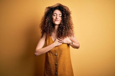 Güzel esmer kadın tatilde. Kıvırcık saçlı, piercing takan, şapkalı ve elbisesi olan elleri göğsünde, gözleri kapalı ve yüzünde minnet ifadesi olan bir kadın. Sağlık kavramı.