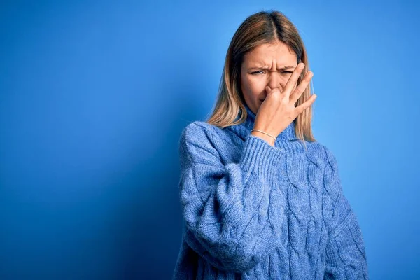 若い美しいブロンドの女性は 青い隔離された背景の上に冬のウールのセーターを着て何か臭いと嫌な 耐え難い匂いを嗅ぎ 鼻の上に指で息を保持します 匂いが悪い — ストック写真