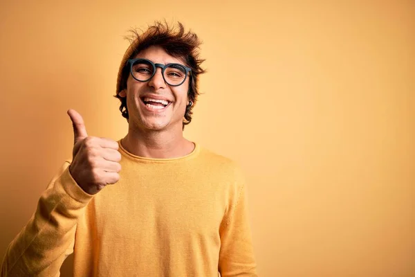 孤立した黄色の背景にカジュアルなTシャツと眼鏡を身に着けている若いハンサムな男は手で幸せな親指アップジェスチャーを行います 成功を示すカメラを見て表現を承認する — ストック写真