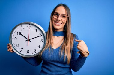 Mavi gözlü genç bir kadın, izole edilmiş arka planda büyük bir saat tutuyor. Şaşırmış yüz ifadesi kendini gösteriyor.