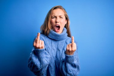 Young beautiful blonde woman wearing casual turtleneck sweater over blue background Showing middle finger doing fuck you bad expression, provocation and rude attitude. Screaming excited