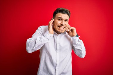 Mavi gözlü, zarif gömlekli genç bir iş adamı kırmızı izole arka planda duruyor. Yüksek sesli müzik sesi için kulakları parmaklarıyla kapatıyor. Sağır kavramı.