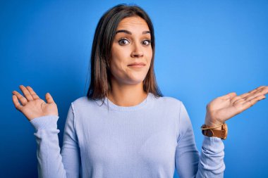 Genç, güzel, esmer bir kadın mavi arka planda dikilen sıradan bir süveter giyiyor. Kolları ve elleri havada, kafası karışmış bir ifade takınıyor. Şüphe kavramı.