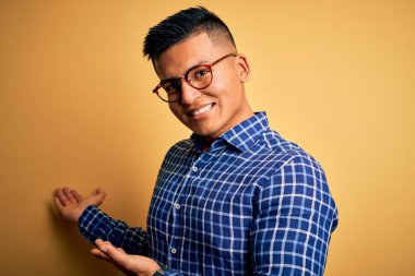 Sarı arka plan üzerine günlük gömlek ve gözlük takan genç, yakışıklı Latin adam gülümseyerek doğal bir şekilde içeri girmeye davet ediyor.