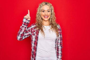 Genç, sarışın, sıradan bir gömlek giyen, kırmızı arka planda duran, bir numaralı parmağını gösterip gülümseyen, kendinden emin ve mutlu bir kadın..