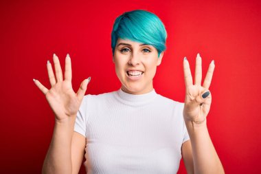 Mavi saçlı, kırmızı arka planda günlük tişört giyen genç ve güzel bir kadın 8 numaralı parmaklarıyla gülümseyerek kendinden emin ve mutlu bir şekilde onu işaret ediyor..