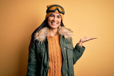 Kış havası için kayak gözlüğü ve palto takan genç ve güzel bir İspanyol kadın neşeli bir şekilde sunum yapıyor ve avuç içi kameraya bakıyor..