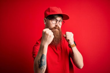 Gözlüklü yakışıklı teslimatçı ve izole edilmiş arka planda kırmızı şapkalı. Yumruk savunma hareketleriyle dövüşmeye hazır. Kızgın ve üzgün yüzlü.