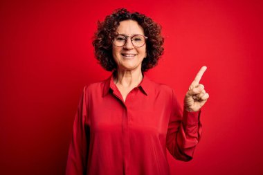 Orta yaşlı, kıvırcık saçlı, rahat gömlekli, kırmızı arka planda gözlüklü, yüzünde kocaman bir gülümseme olan güzel bir kadın..