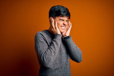 İzole edilmiş turuncu arka planda dikilen sıradan süveter giyen genç yakışıklı yüksek sesli müzik sesi için parmaklarıyla kulaklarını kapatıyor. Sağır kavramı.