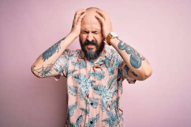 Sakalı ve dövmesi olan yakışıklı kel bir adam pembe arka planda sıradan bir çiçek gömleği giyiyor. Ağrı ve migrenden dolayı çaresiz ve stresli. Ellerini başının üstüne koy..