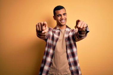 Genç, yakışıklı, Afro-Amerikalı, günlük gömlekler giyen, sarı arka planda duran, sana ve kameraya parmaklarıyla gülümseyen, pozitif ve neşeli bir adam.