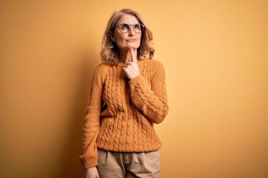 Orta yaşlı güzel sarışın kadın sarı arka planda günlük süveter ve gözlük takıyor. Şüphe üzerine yoğunlaşmış, çenesinde parmak olan ve kafasını kaldırıp merak eden biri.