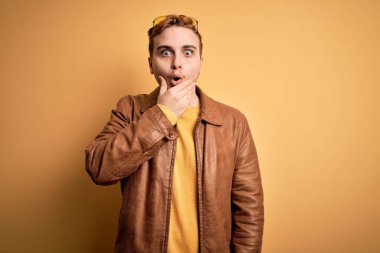 İzole edilmiş sarı arka plan üzerine sıradan deri ceket giyen genç, yakışıklı kızıl saçlı adam elleri çenesinde olan inançsızlıktan, şaşkınlıktan ve şaşkınlıktan büyülenmiş görünüyor.