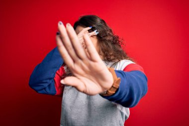 Kıvırcık saçlı genç bir kadın izole edilmiş kırmızı arka plan üzerine günlük eşofman giyip gözleri ellerle kaplıyor ve üzgün ve korku ifadeleriyle jest yapıyor. Utanç verici ve olumsuz bir kavram..
