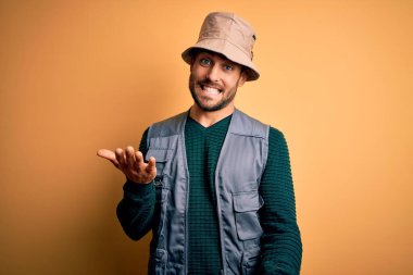 Tatilde sakallı yakışıklı turist sarı arka planda kaşif şapkası takıyor. Kolları açık gülümsüyor. Dostça karşılama, olumlu ve kendinden emin selamlar.