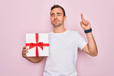 Mavi gözlü genç adam pembe arka planda doğum günü hediyesini tutarken bir fikir ya da soru işareti ile şaşırdı.