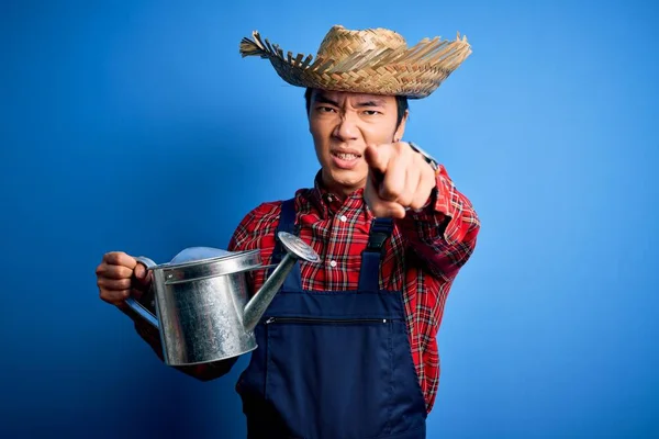Young handsome chinese farmer man wearing apron and straw hat holding watering can pointing with finger to the camera and to you, hand sign, positive and confident gesture from the front