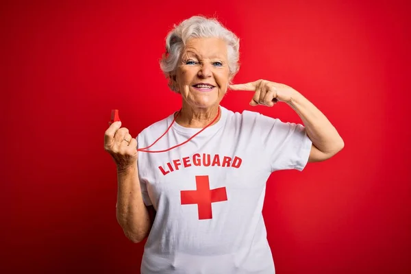 赤い十字のTシャツを着た上級の美しい白髪のライフガードの女性は 明るい表情と指の歯と口で指差す笛を使用しています 歯科衛生の概念 — ストック写真