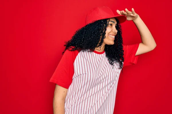Young african american curly sportswoman wearing baseball cap and striped t-shirt very happy and smiling looking far away with hand over head. Searching concept.