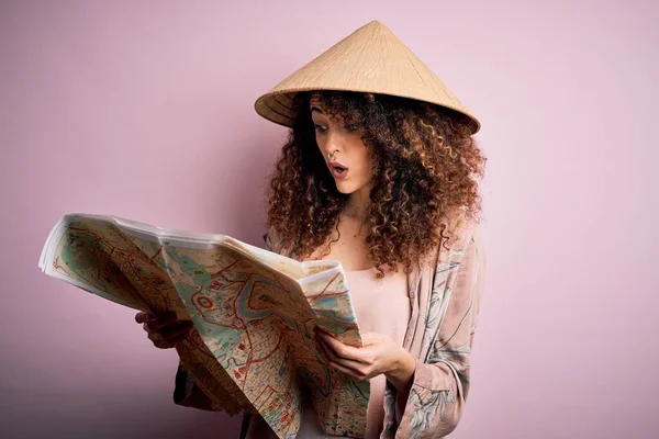 Young beautiful tourist woman with curly hair and piercing wearing asian hat holding city map scared in shock with a surprise face, afraid and excited with fear expression