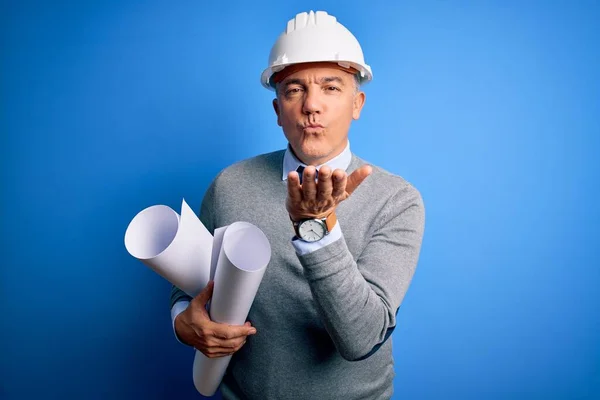 Middle age handsome grey-haired architect man wearing safety helmet holding blueprints looking at the camera blowing a kiss with hand on air being lovely and sexy. Love expression.